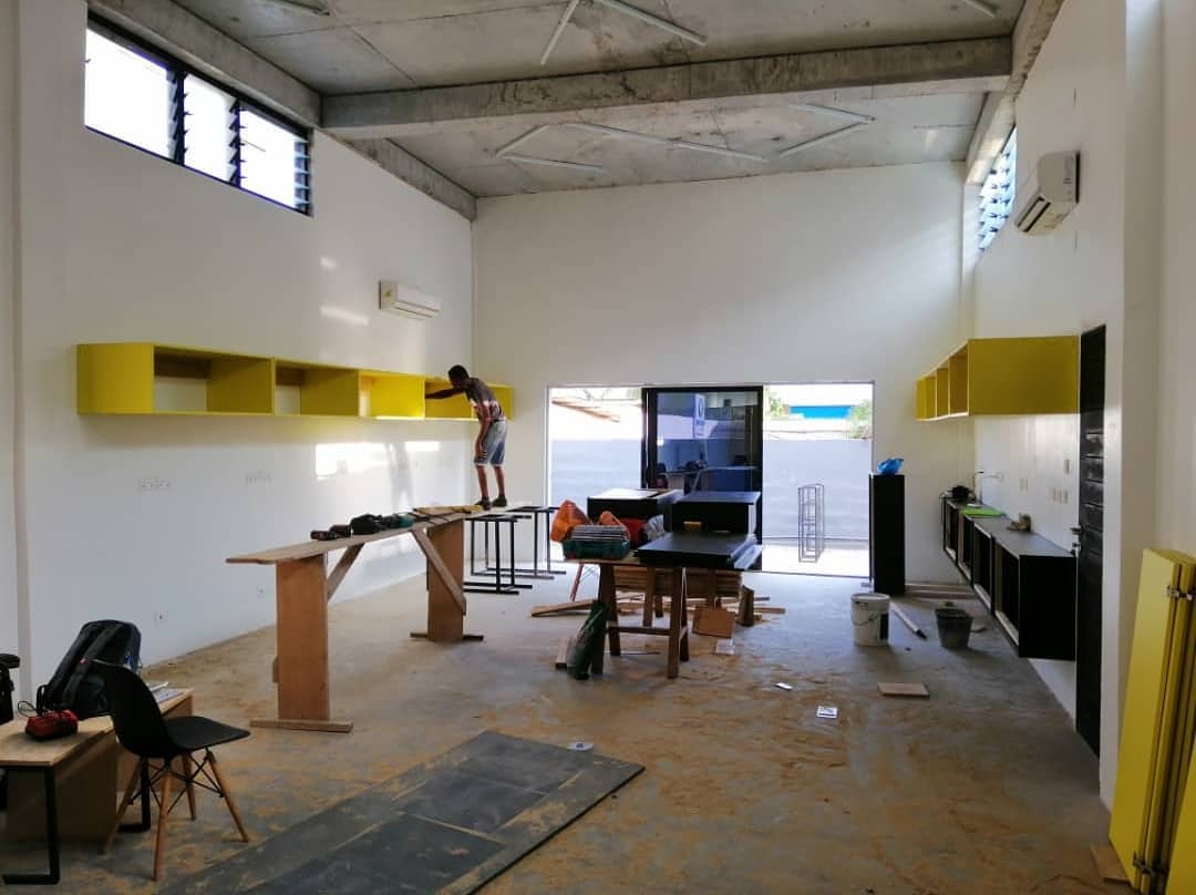 3AAP interior fit-out in progress: a tall white room under an exposed concrete ceiling and steel trusses, a worker standing on a makeshift plywood bench to fix a run of yellow overhead cabinets, black base units along the right wall and an open door to the courtyard at the far end