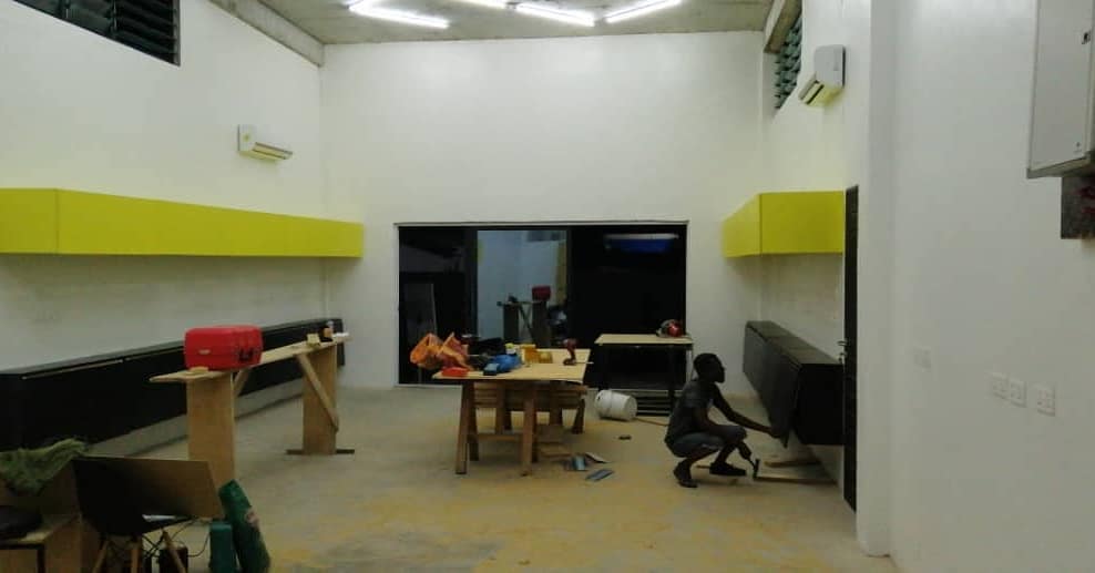 3AAP — the same room after dark under fluorescent battens: the yellow overhead cabinets now running the length of both walls above their black base units, a worker crouched at the right-hand counter, the makeshift benches still standing in the middle of the floor