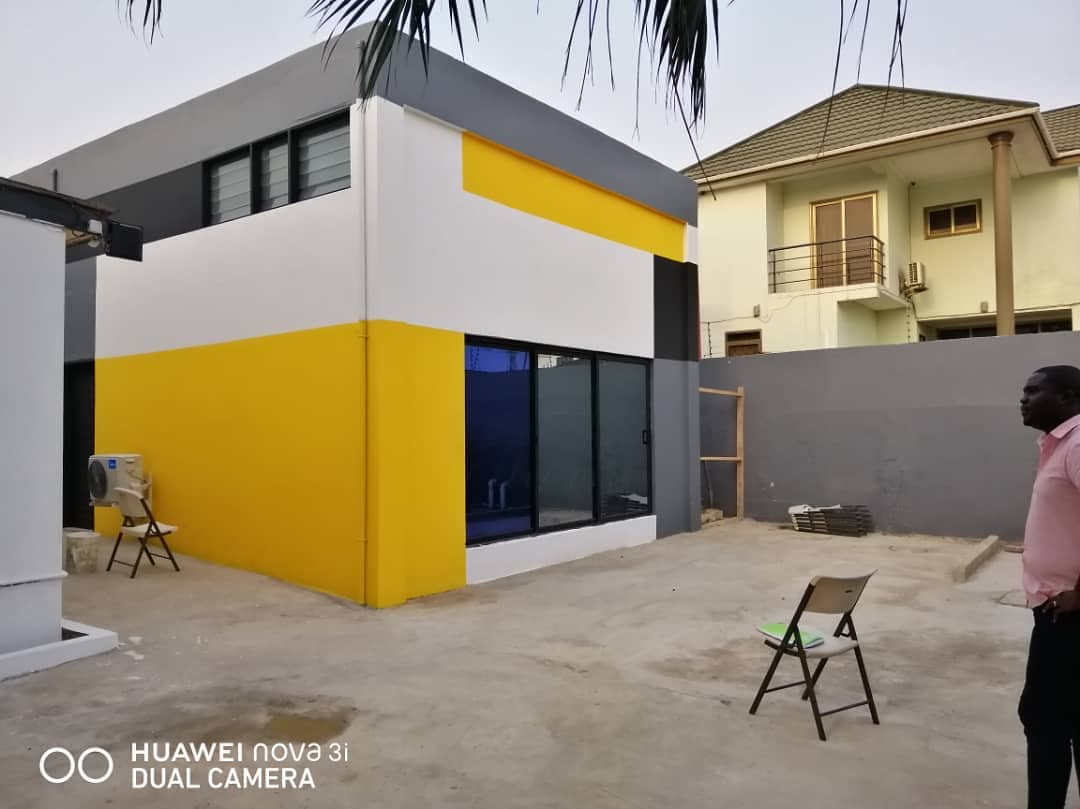 3AAP — the corner of the building from the yard: a yellow ground floor, white banding and a grey upper volume, with dark-framed glazing wrapping the corner and a neighbouring house beyond the boundary wall
