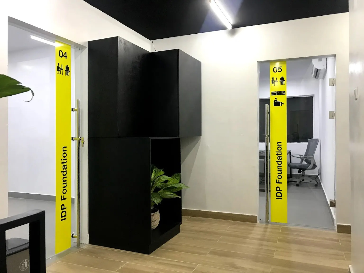 5AAP built signage in the IDP Foundation office: full-height yellow bands on two glass doors reading 04 and 05 with meeting-room pictograms and IDP Foundation set vertically in black, a stacked black cube divider holding a potted peace lily between them