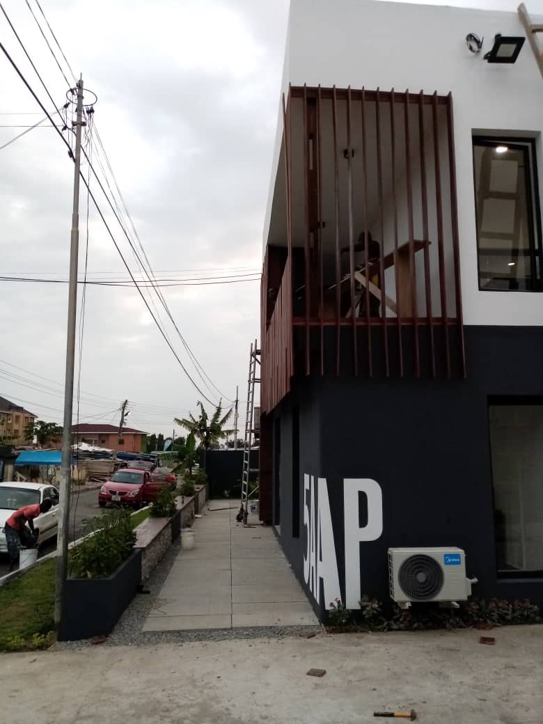 5AAP — the street elevation with the corner wordmark painted and the batten screen in place, a worker on the first-floor balcony