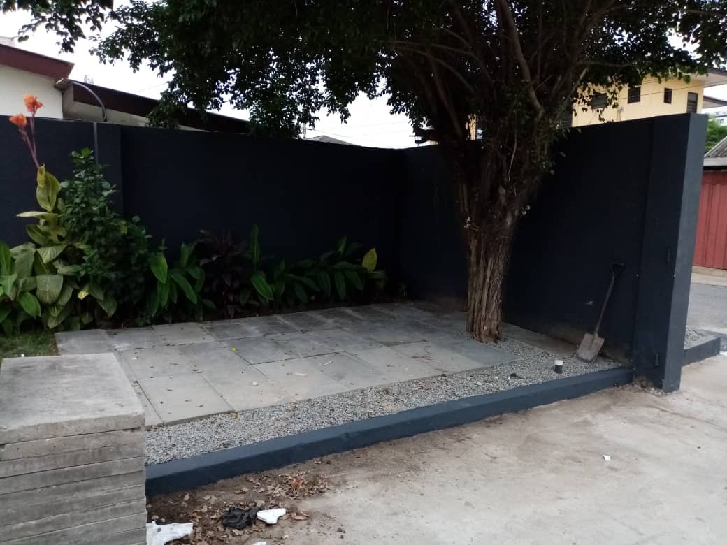 5AAP — the courtyard: a black boundary wall with a planted bed at its base, an existing tree rising through newly laid paving and a spade left against the wall