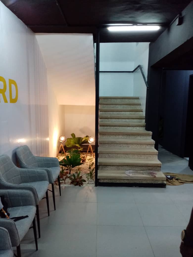 5AAP — the waiting lounge: quilted grey armchairs beside an interior planted bed lit by tripod lamps, yellow wall lettering running out of frame and timber stairs rising at right