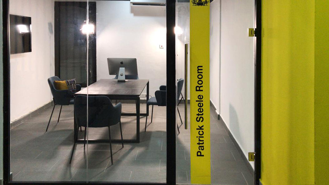 5AAP — the Patrick Steele Room seen through its full-height glass partition: dark table, blue chairs and a computer at the far end