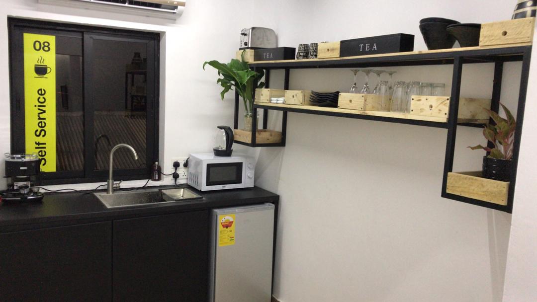 5AAP — the pantry: black counter, stainless sink, microwave and kettle beneath a window vinylled '08 Self Service', with open shelving holding tea crates and mugs