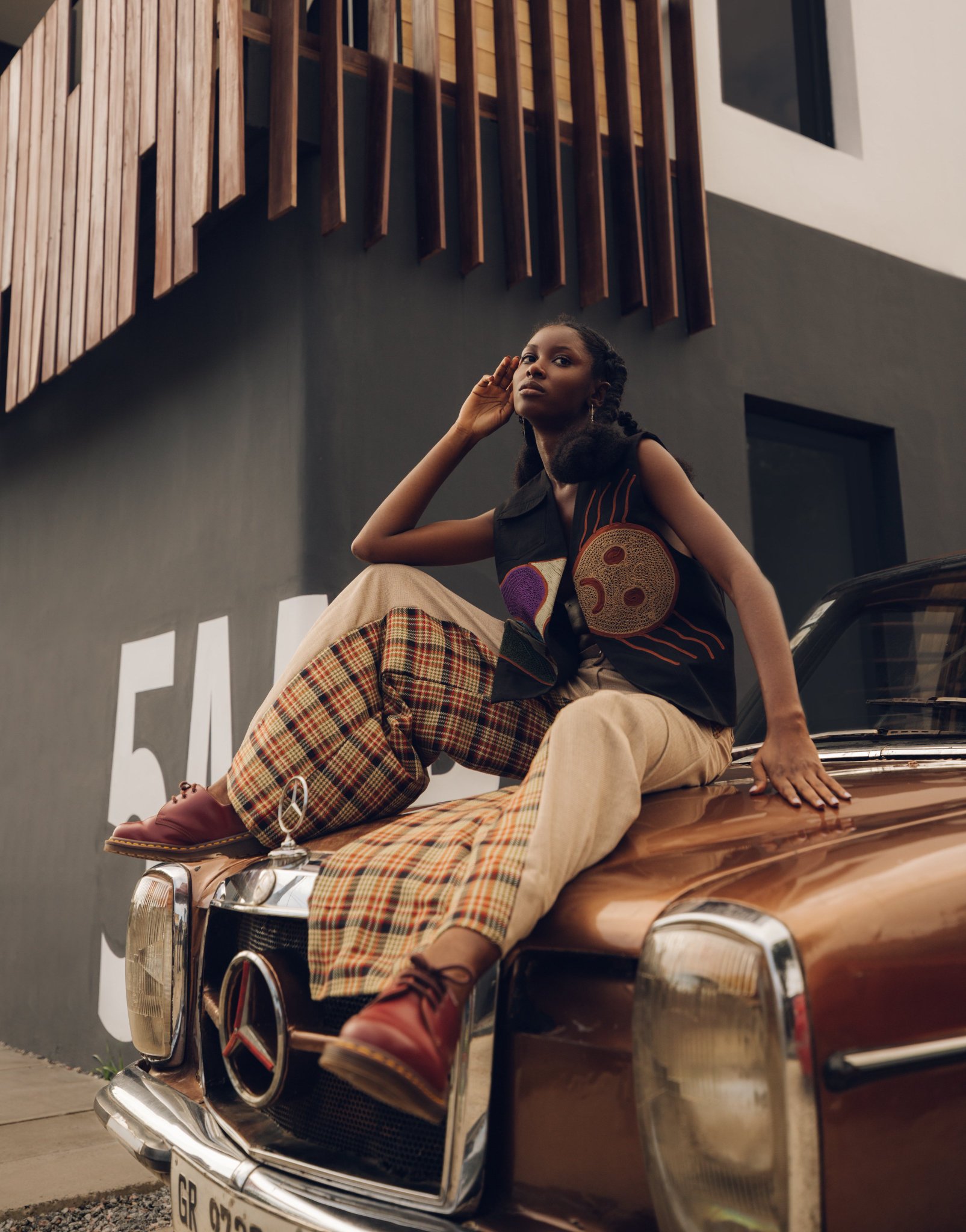 5AAP lookbook frame: a model seated on the bonnet of a copper-brown vintage Mercedes before a dark grey wall carrying oversized white lettering and timber louvres above, in an embroidered black vest, checked and cream trousers and red leather shoes