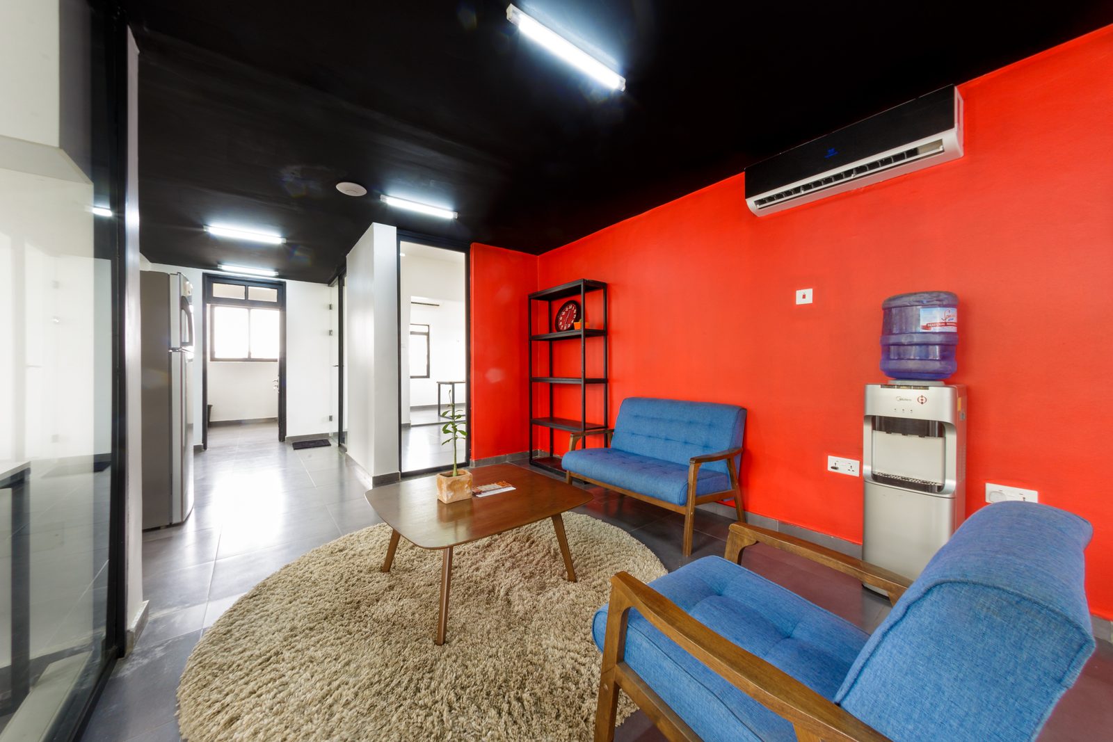AFG - The lounge from the red wall: black open shelving, a water dispenser and the blue sofa grouped on a circular rug around a plywood coffee table.