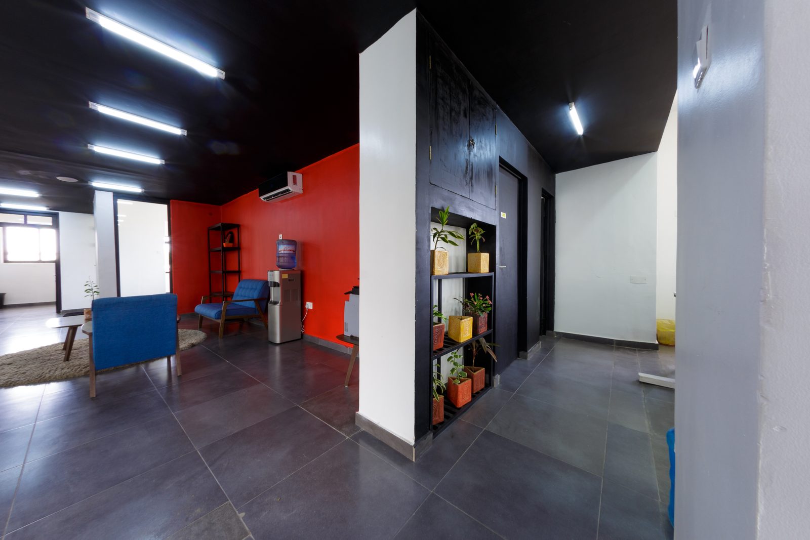 AFG - A black shelving unit loaded with plants and books anchoring the corner of the lounge, the corridor opening away to the right.