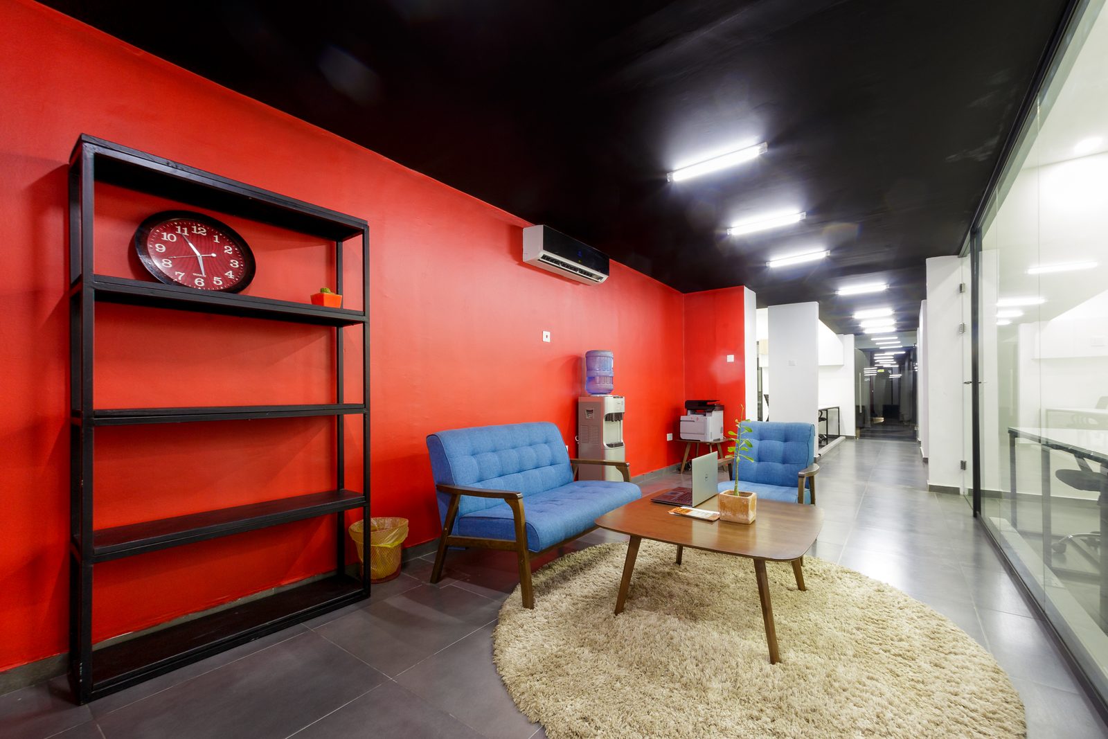 AFG - The red wall of the lounge with its black open shelving and a round wall clock, the sofa and coffee table on the rug below.