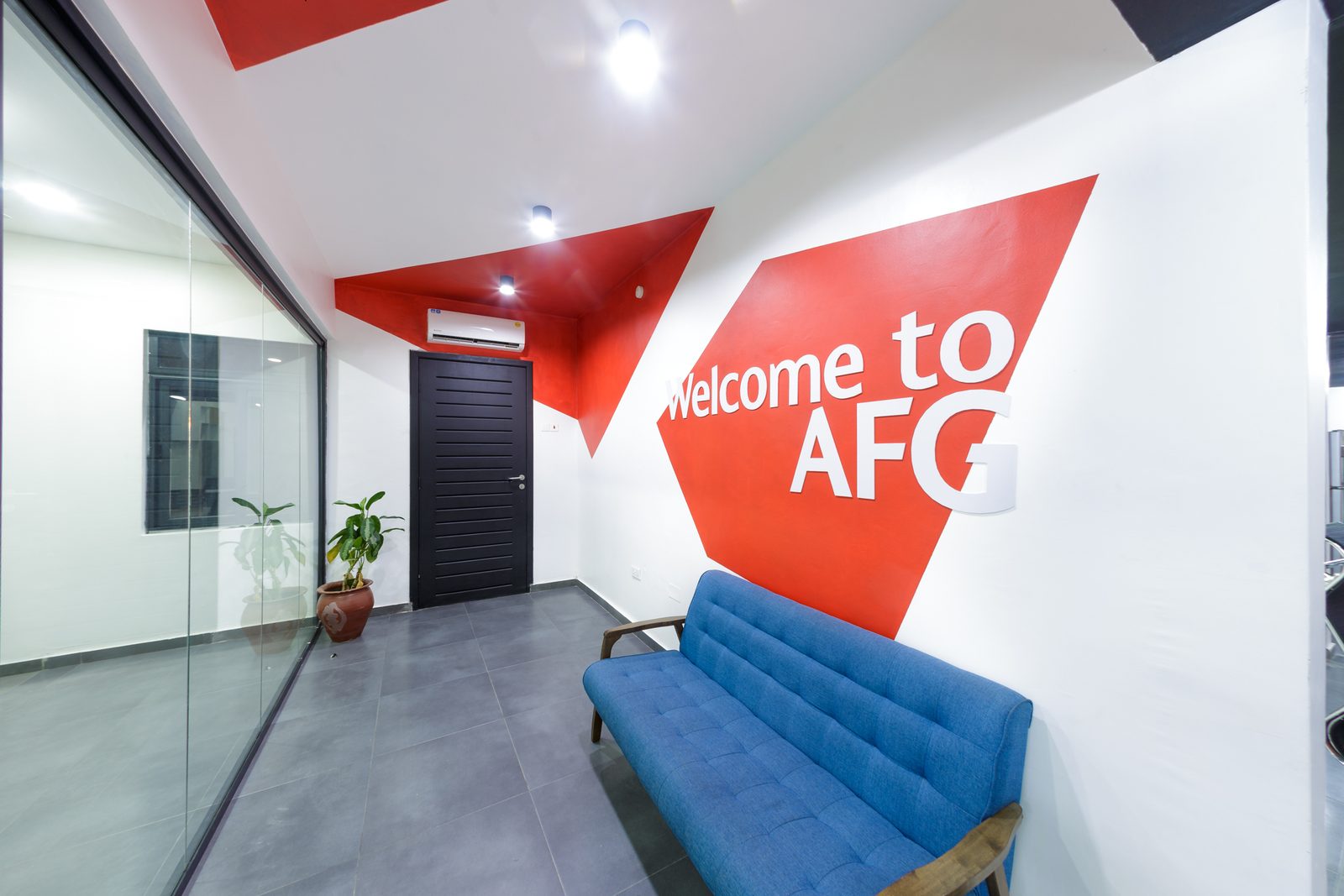 AFG - The same lounge corner from further along the corridor, the Welcome to AFG graphic filling the wall above a two-seat blue sofa under a black ceiling.
