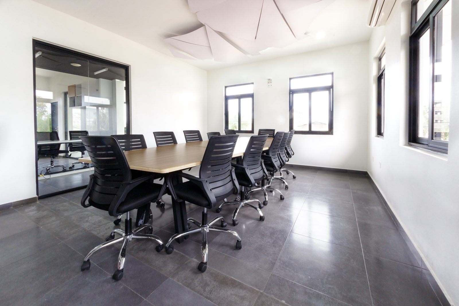 AFG - The boardroom from the head of the table: a long plywood-topped conference table with black mesh chairs, the faceted white acoustic ceiling above and the glazed wall back to the lounge.