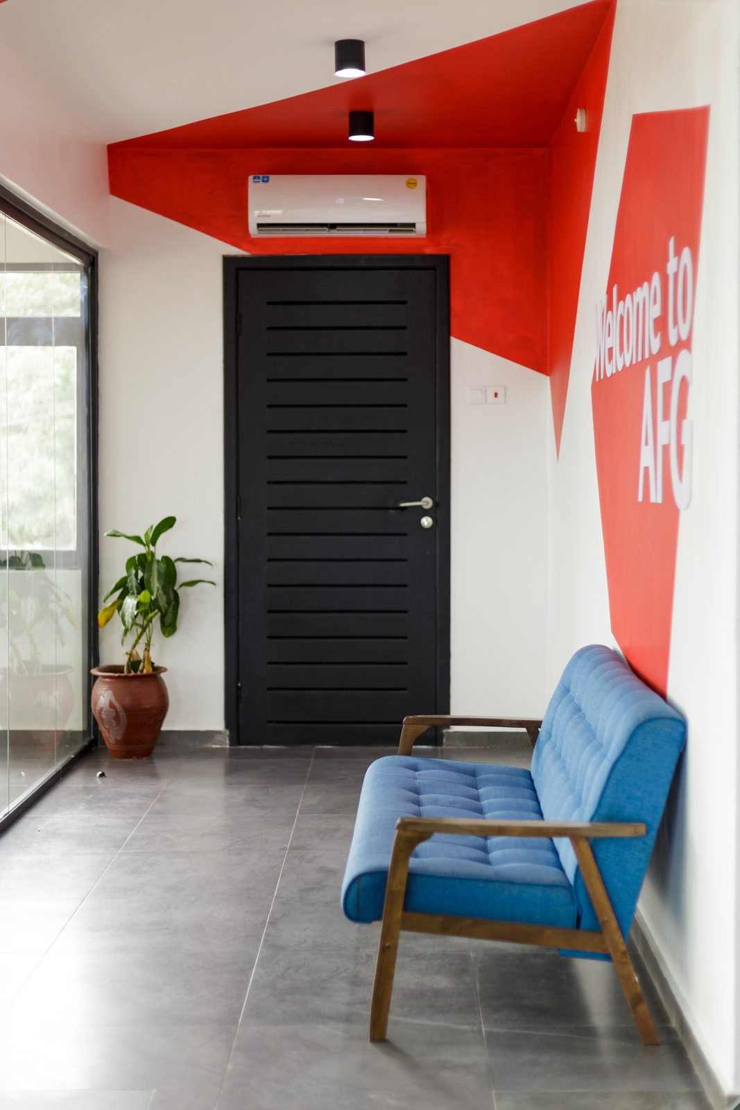 AFG - Portrait view of the entrance wall, the black door and wall-mounted air-conditioning unit framed by angular red graphics and a train of downlights on the black ceiling.