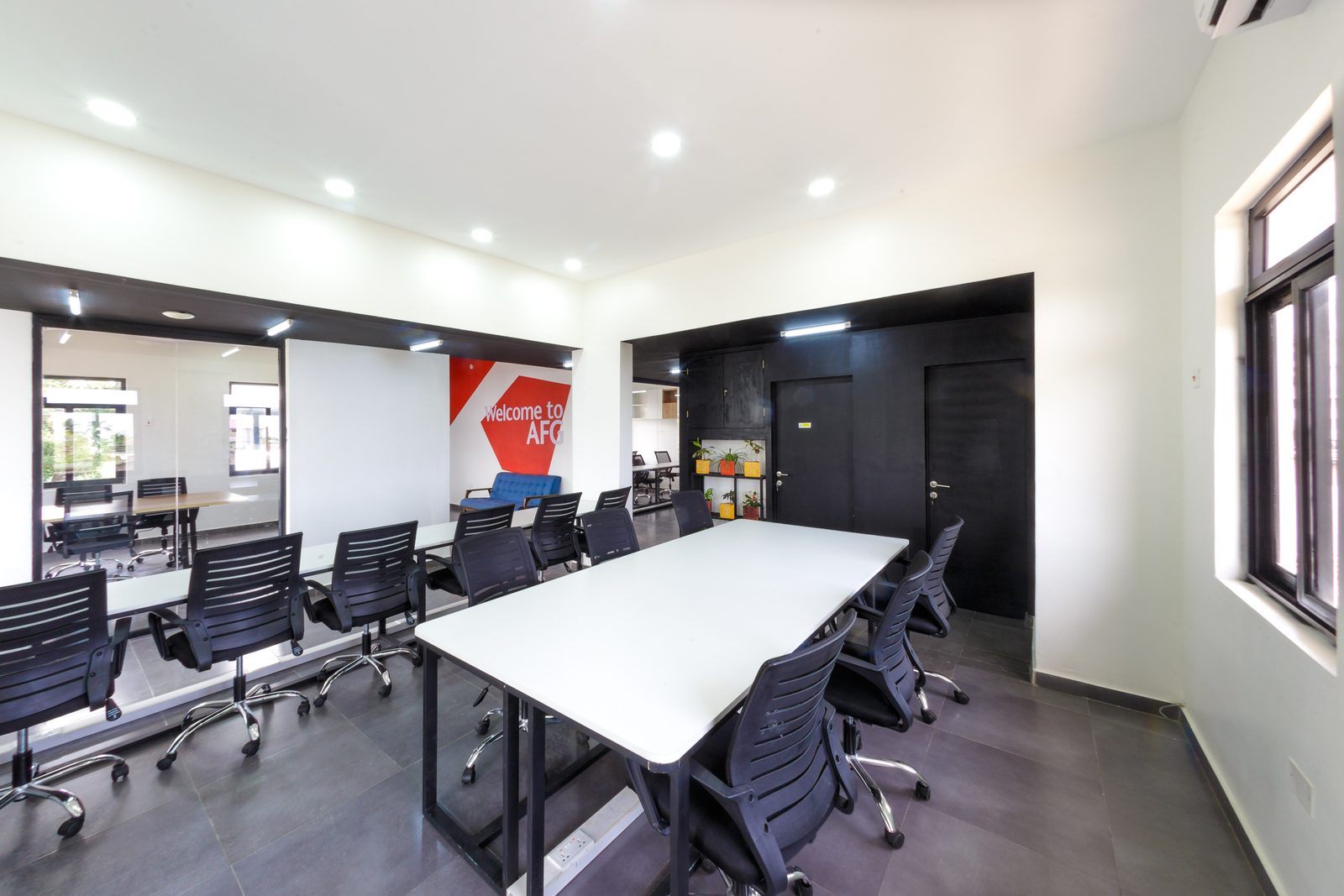 AFG - The open workspace: white-topped bench desks with black mesh chairs under a white ceiling of downlights, the black-painted zone and red graphic wall at the far end.