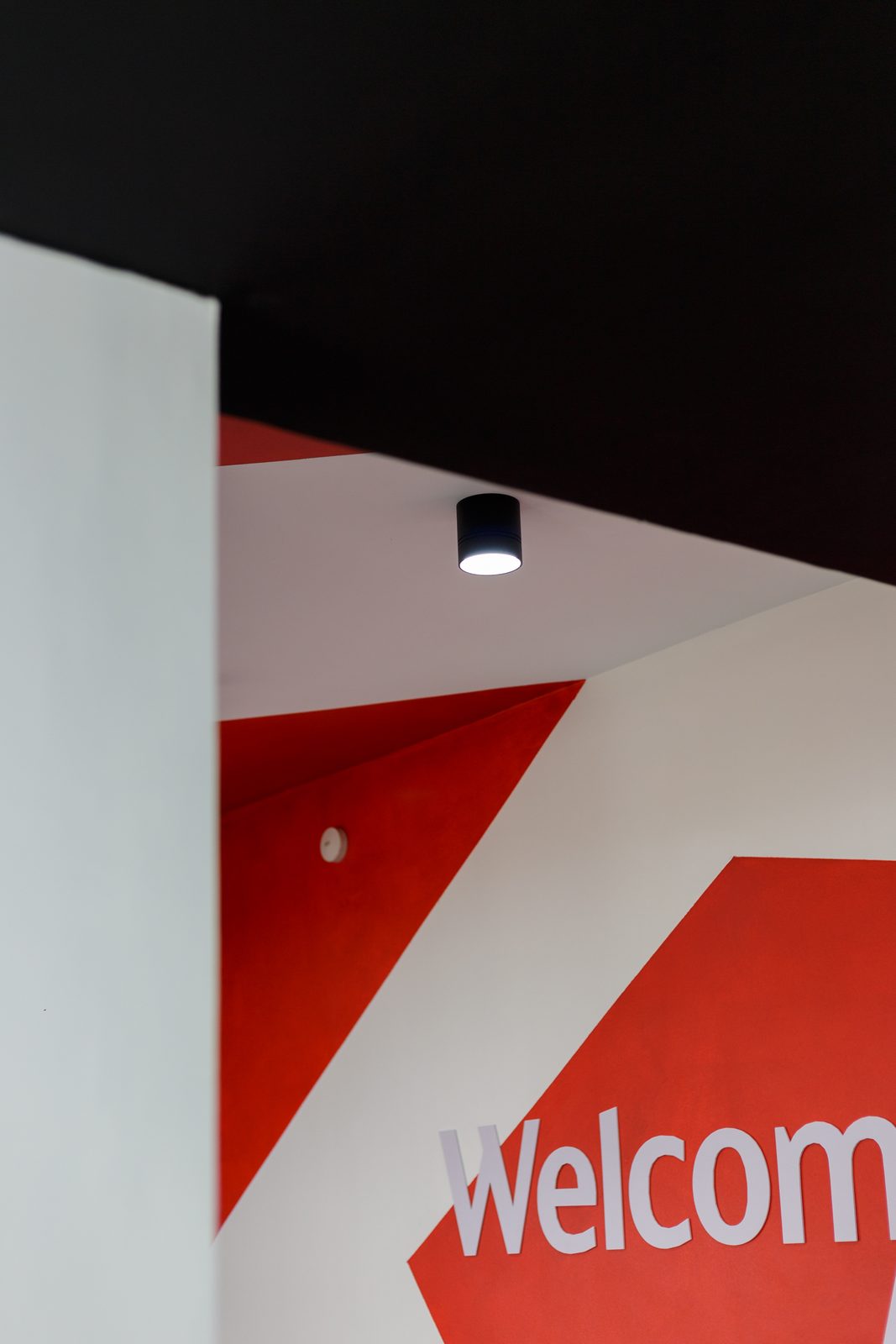 AFG - A close crop of the graphic wall, the red Welcome lettering cut by a white column and a single surface-mounted spotlight.