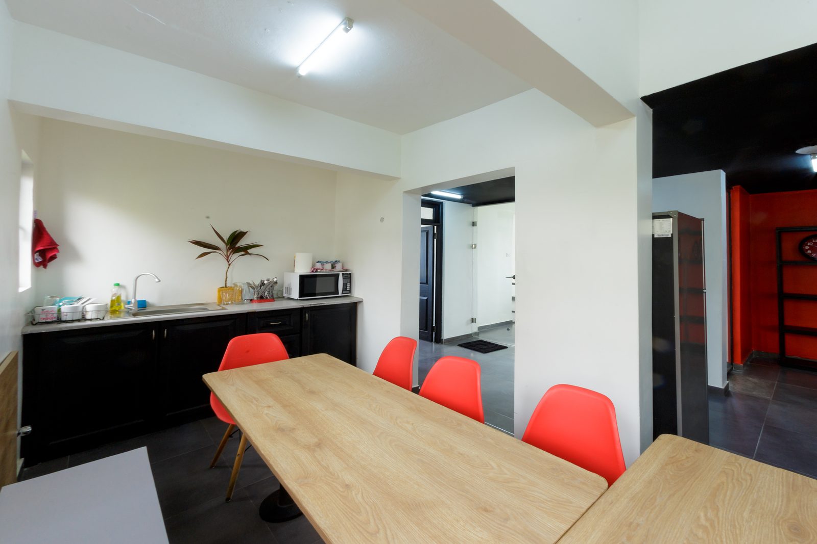 AFG - The pantry: black base cabinets and a plywood counter with sink and microwave, a long plywood table with red moulded chairs in front.