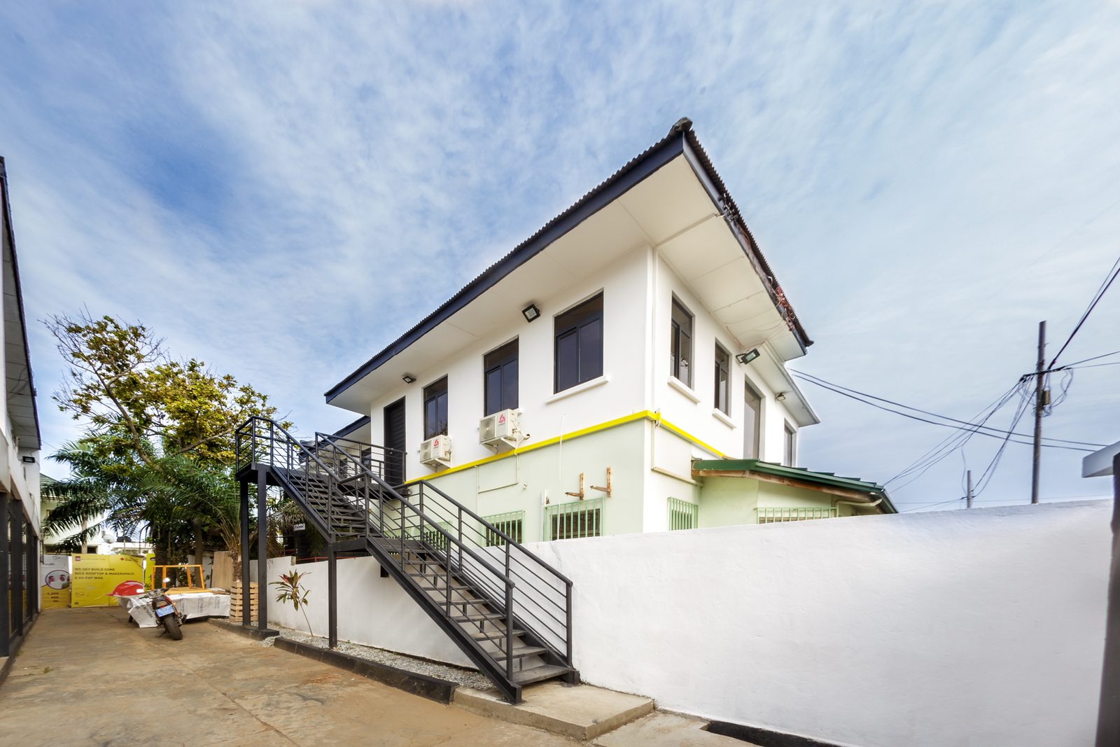 AFG - The building from the yard: a white two-storey block with ochre banding, an external black steel stair and a rendered boundary wall.