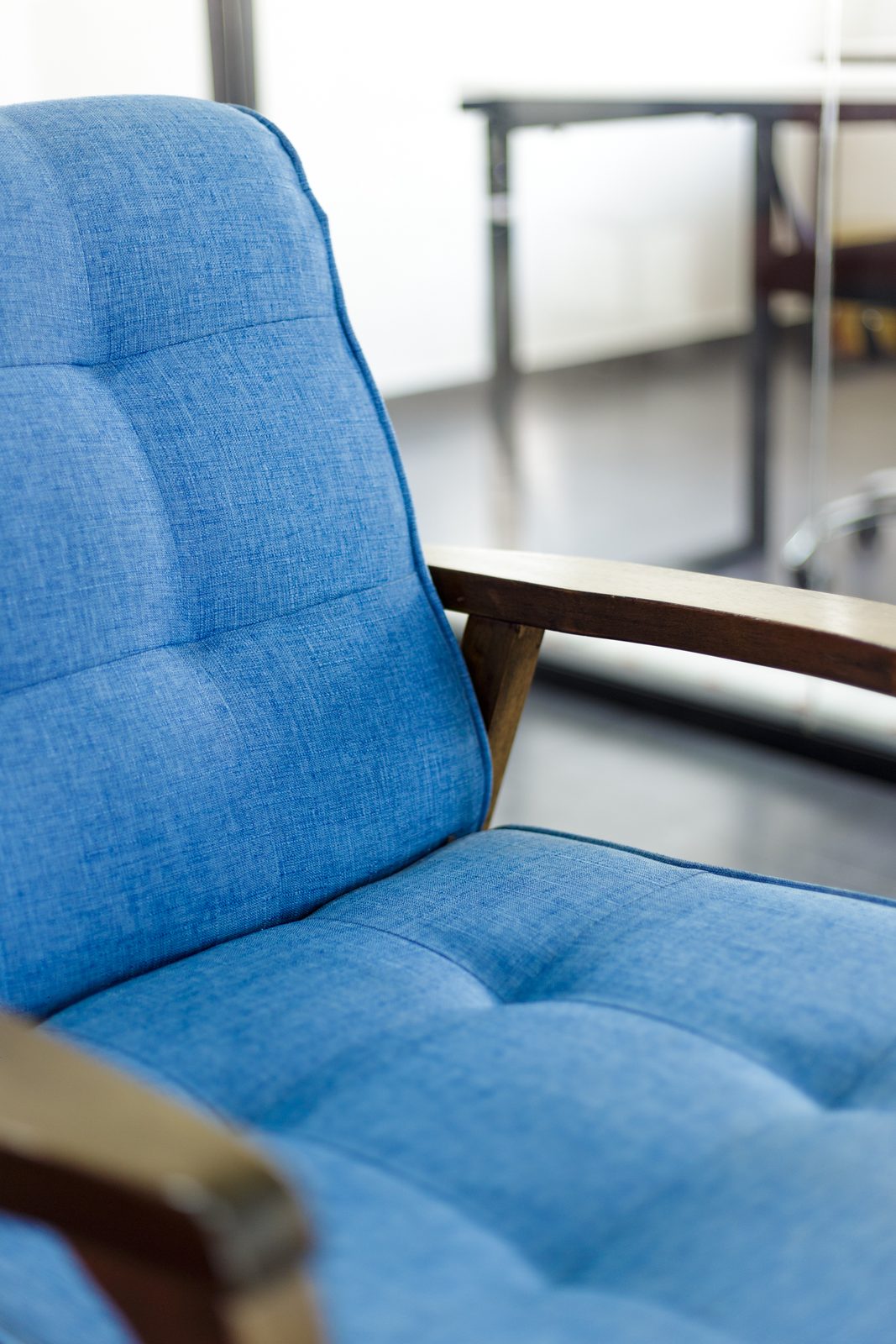 AFG - Tight detail of the blue tufted upholstery and timber frame of the lounge chair, the open plan out of focus beyond.