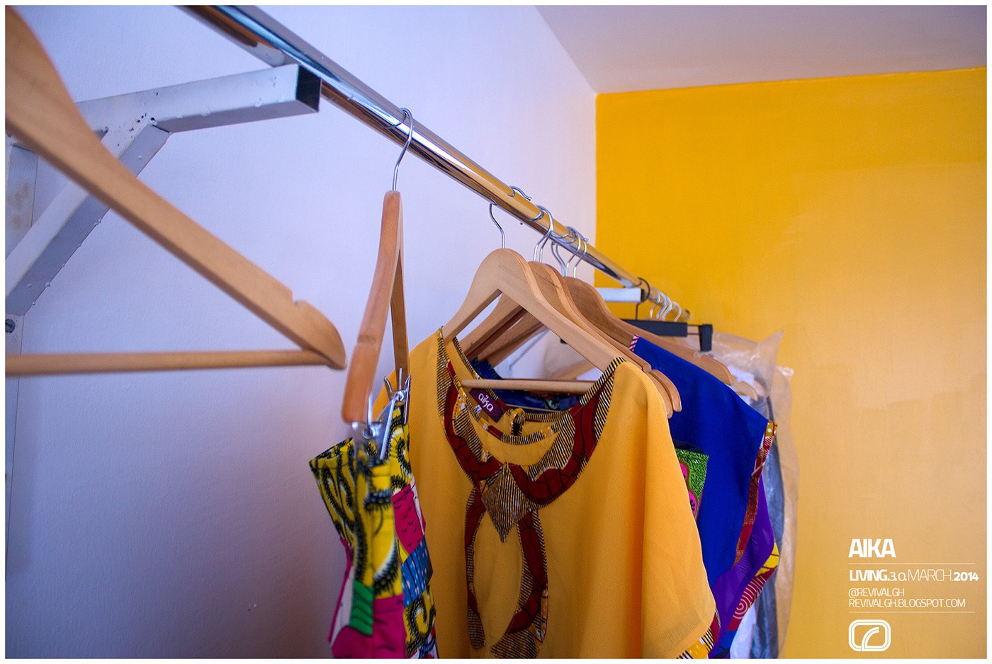 Photograph, close interior view: wooden hangers carrying a printed blouse and a gold top on a chrome wall rail, white wall with a yellow panel behind, AIKA wordmark and a small credit block at lower right.