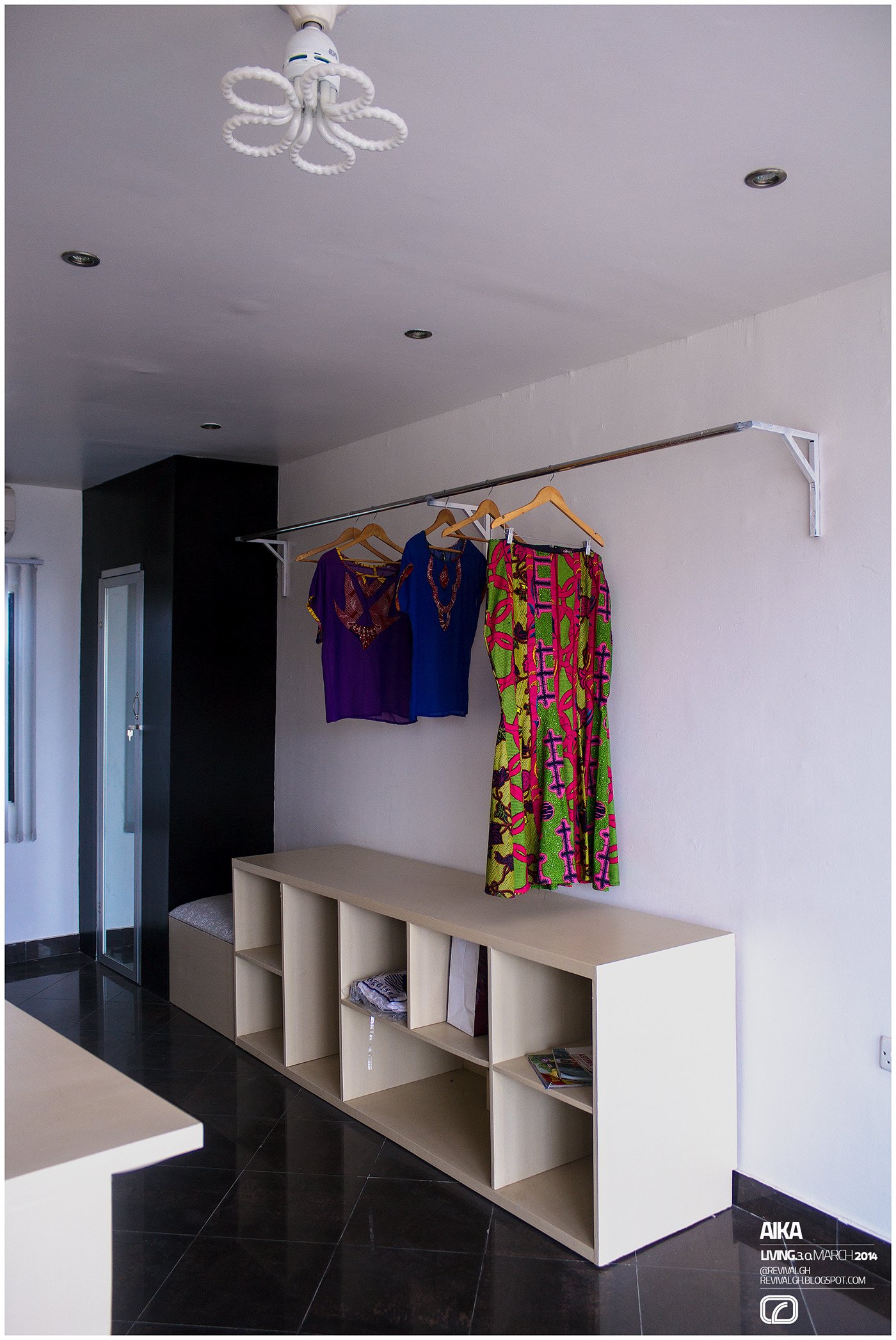 Photograph, wide view of the selling floor: rails of purple, blue and pink wax-print garments above a low white open-cubby bench, glossy black tiled floor, white ceiling fan and downlights, black-clad column and doorway at left.