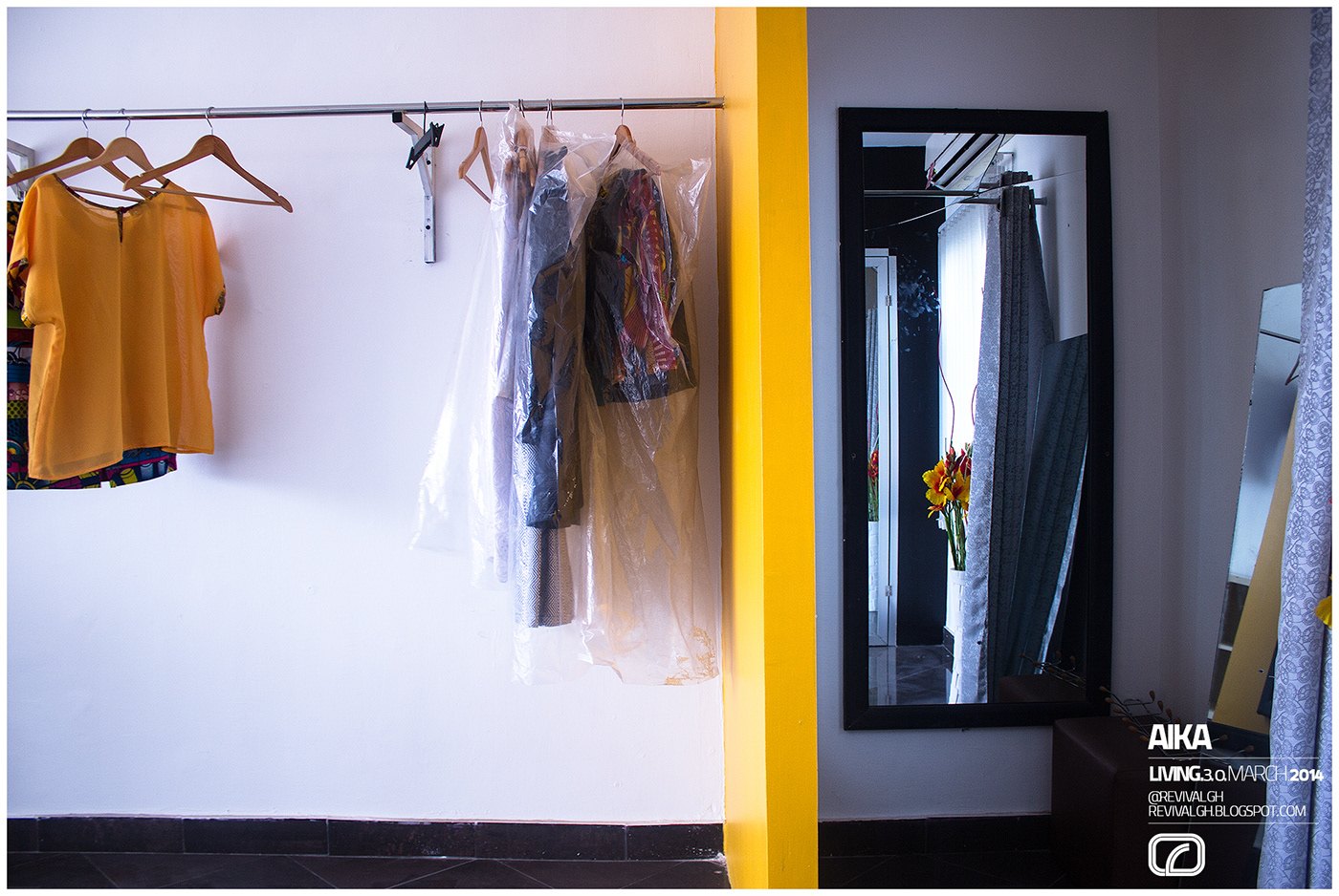 Photograph, the mirror end of the shop: a mustard tee and covered garments hung on a wall rail, a yellow pier, tall black-framed mirror reflecting the room, dark floor and an AIKA credit block at lower right.