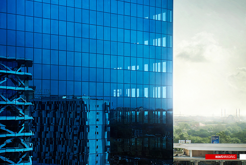 A blue glass curtain wall seen close along its corner, filling the left two-thirds of the frame with a grid of square reflective panes. The lower half of the glazing works as a mirror: it returns a neighbouring facade clad in an irregular checkerboard of black, white and grey panels together with a rooftop sign whose letters run backwards in the glass. Past the right edge of the building the frame opens onto a washed-out white sky, a low hazy treeline and a single blue billboard, and a red rectangular RDVSIMAGING badge sits in the bottom-right corner.
