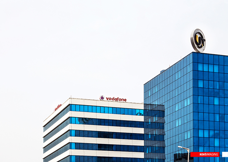 Two office rooftops cropped against an almost featureless white sky. On the left a white banded building with horizontal strips of blue glazing carries the Vodafone wordmark in red lowercase on its parapet, with a second red sign running away from the camera along the same roofline; on the right a taller tower of blue glass carries a circular gold-and-black dimensional monogram at its corner. The head of a street lamp intrudes at the bottom edge. Red RDVSIMAGING badge, bottom right.