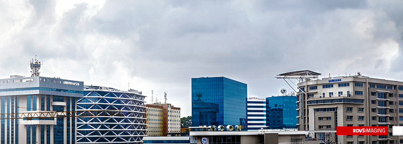 A very wide, low strip of skyline under heavy grey cloud: a cylindrical glass atrium braced with white diagrid at the left and a construction crane jib crossing in front of it, beige slab blocks and blue-glass towers packed along the horizon, one saturated blue cube standing out near the centre, and a run of large dimensional rooftop capitals along the low building in the middle distance with a blue shield badge on its corner. Red RDVSIMAGING badge, bottom right.