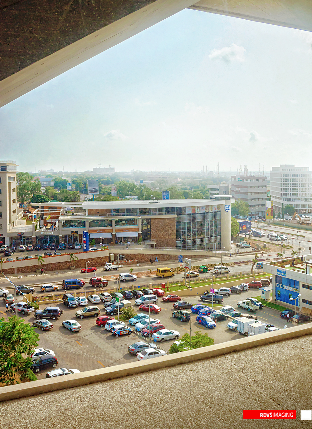 A tall frame looking downward through the wedge-shaped gap between two concrete slabs: the dark underside of a floor crosses the top of the image and a pale parapet closes the bottom, so the view is cut to an irregular opening. Through it sits a multi-storey open car park packed with cars, a low two-storey retail building with a cylindrical corner tower carrying a blue shield badge, a divided road with a yellow minibus, and the flat hazy Accra horizon with industrial chimneys and a distant white office block. Red RDVSIMAGING badge, bottom right.