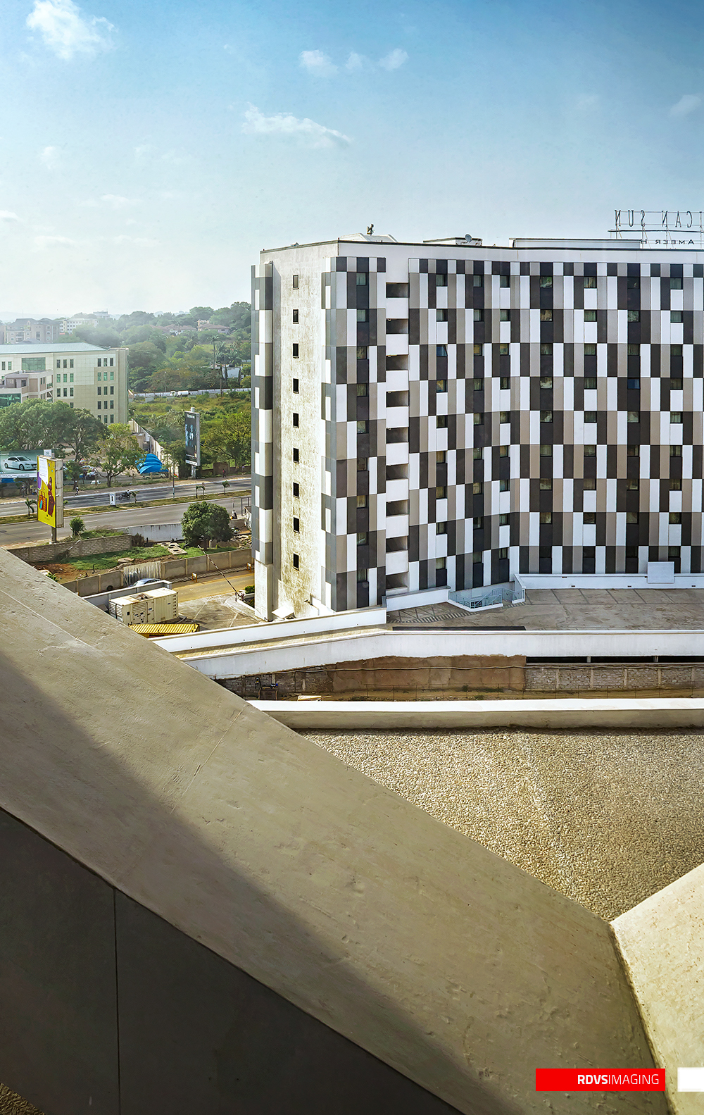 The tallest frame, looking down at a mid-rise building whose two visible faces are clad in an irregular checkerboard of black, white, grey and beige panels with deeply recessed windows. A concrete parapet or stair soffit cuts diagonally across the whole lower half of the image, close to the lens; a service road, a generator set and a yellow billboard sit at the left, a green treeline and low buildings fill the middle distance, and a wide pale blue sky with thin cloud fills the top. A rooftop sign on the checkered building is seen from behind, its letters running the other way. Red RDVSIMAGING badge, bottom right.