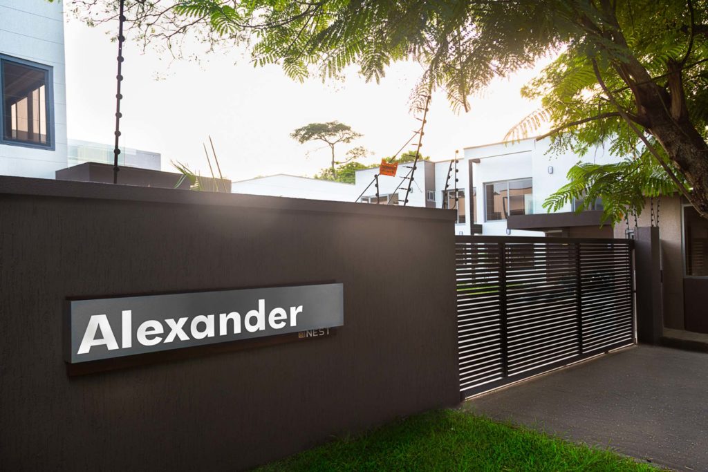 Alexander-Signage in place: a backlit grey Alexander plaque with small NEST lettering beneath, fixed to a dark boundary wall beside a slatted black sliding gate, white houses and a flame tree beyond and low sun flaring across the frame