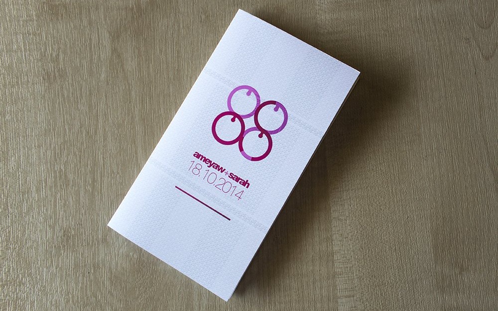 Ameyaw + Sarah — the printed invitation card laid on a wooden table, its Me Ware Wo monogram and the date 18.10.2014 in burgundy on textured white stock