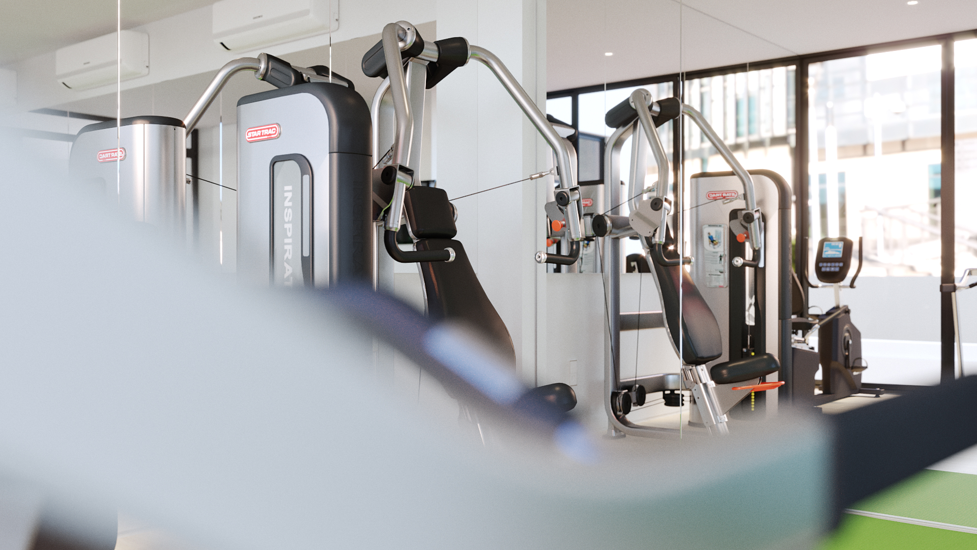 Details — the fitness room, seen past a blurred white rail: a cable training machine and benches against a mirrored wall, full-height windows throwing daylight across the floor