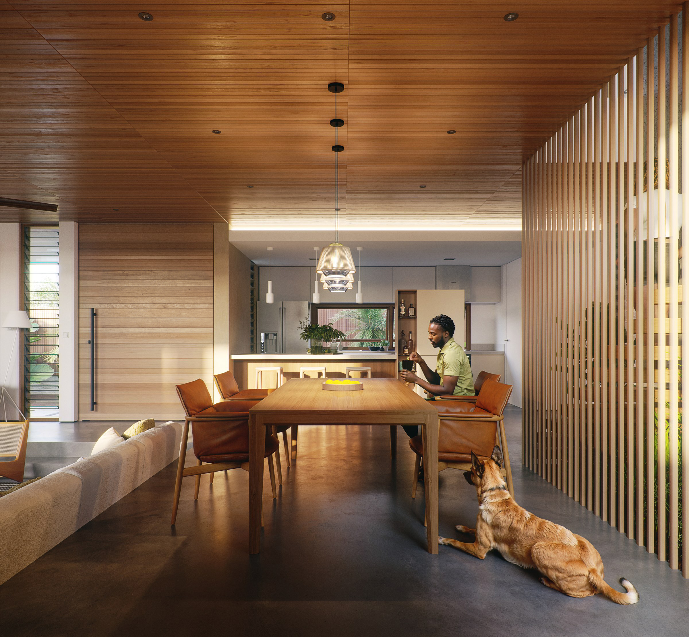 Dining table seen from the living end: a man seated at a timber table in tan leather armchairs beneath a single glass pendant, a red hound resting on the polished floor at his side, the white kitchen and its lit counter behind him and a full-height screen of vertical timber slats to the right.