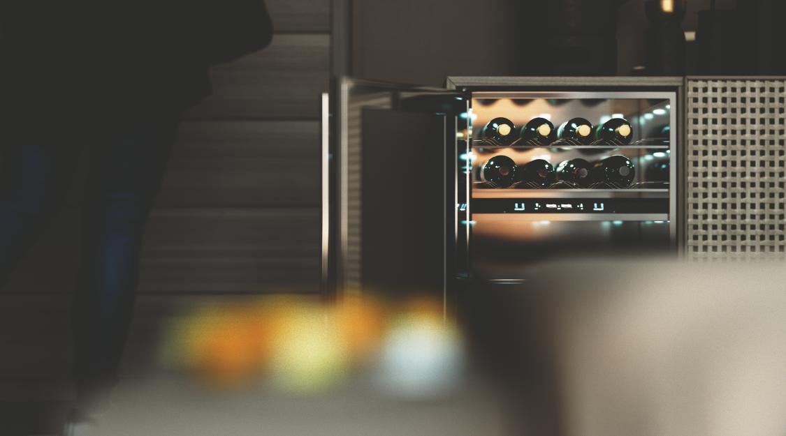 HGG joinery beverage cooler: its glass door open on two rows of bottles under warm interior light, a caned cabinet door to the right and fruit blurred across the foreground.