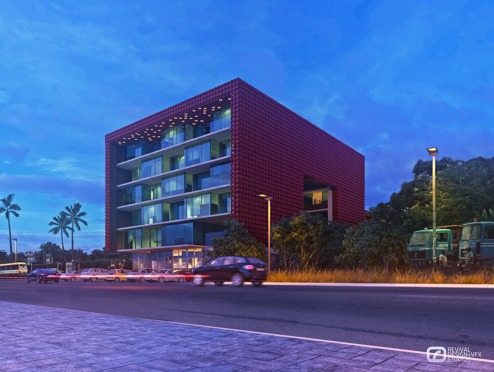 KDMRD at dusk: the red cubic building seen across a wide paved street, its perforated brick-red screen wall glowing against a deep blue sky, glazed balconies lit on the upper floors, palm trees and traffic passing along the roadside.