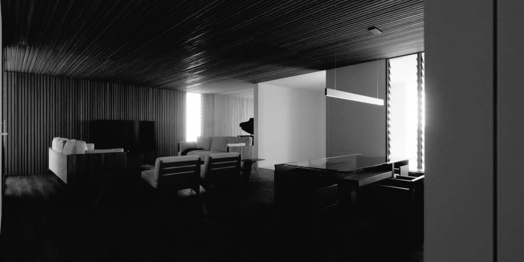 Kuma Residence in monochrome: pale sofas and armchairs against a fluted timber wall under a dark slatted ceiling, the dining table to the right beneath a linear pendant and bright full-height glass beyond