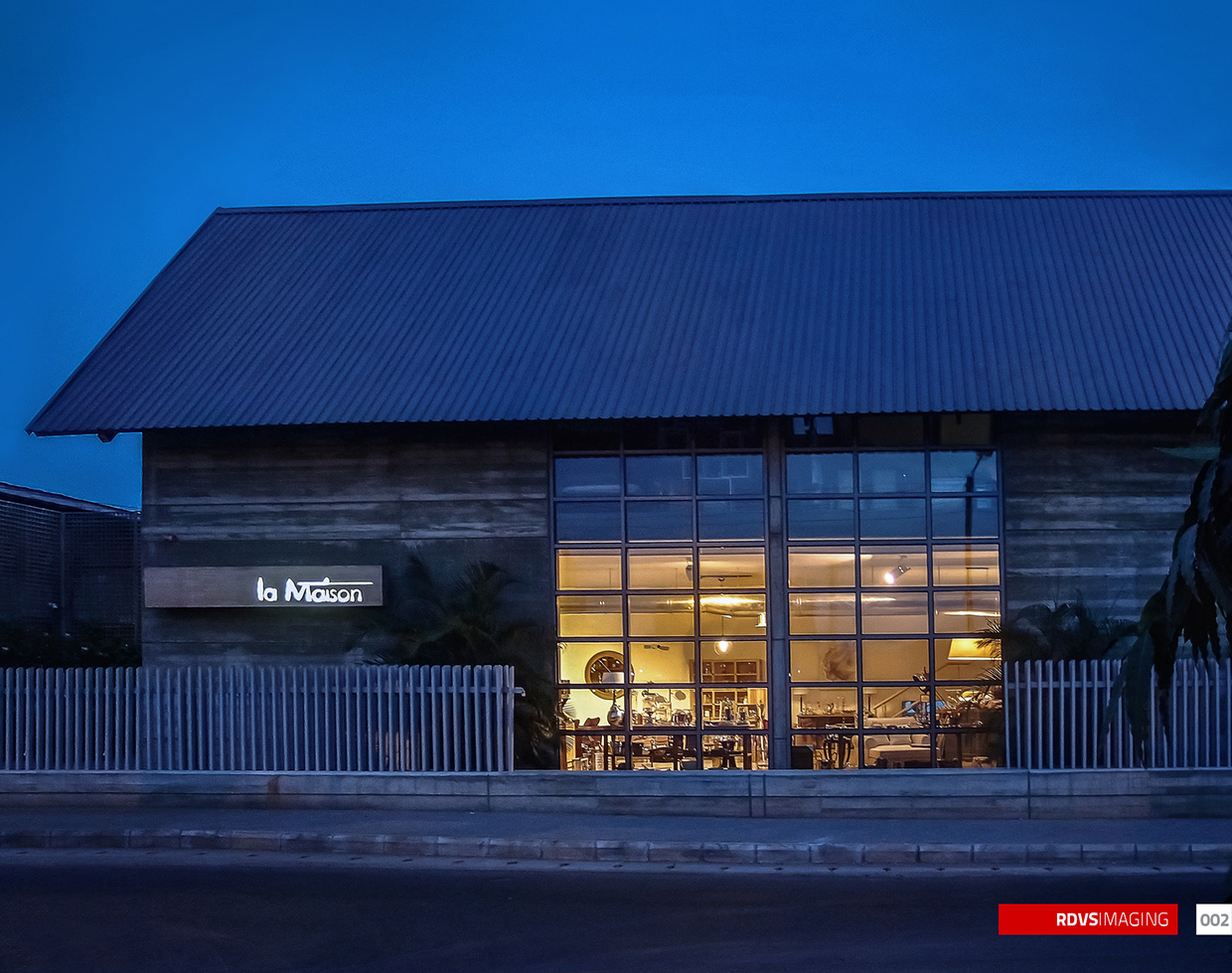 A single-storey building seen square on across a road at dusk: a steep corrugated steel gable roof spans the whole width against a clear deep blue evening sky, and below it the wall is banded horizontally in earth-toned board-marked concrete. The right half of the facade is a full-height grid of steel-framed glazing lit warm gold from inside, through which chairs, tables, a shaded lamp and a large round wall clock are visible. To the left of the glazing a dark blade sign carries the words la Maison in white, with the first word small and the second larger. A pale grey fence of vertical bars on a low concrete plinth runs the length of the frontage, a palm leans into the frame beside the glazing, and asphalt and a stone kerb fill the foreground. Red RDVSIMAGING badge and a white tag numbered 002 at the bottom right.