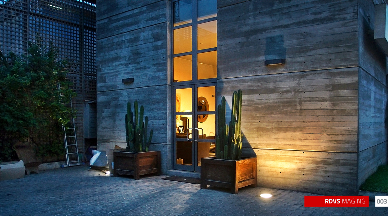 The courtyard corner of the same building at dusk, with the banded earth-toned wall filling the right two-thirds. A tall narrow steel-framed glazed door sits at the centre glowing warm from within, flanked by two timber planters holding clusters of tall columnar cacti. An uplighter washes the wall at the right in a warm fan and a ground spotlight lights the paving, which is laid in grey herringbone blocks. At the left a perforated lattice screen wall rises behind a leafy tree, with an aluminium ladder leaning against it and a board lying on the ground. Two small dark wall lights are fixed high on the concrete. Red RDVSIMAGING badge and a white tag numbered 003 at the bottom right.
