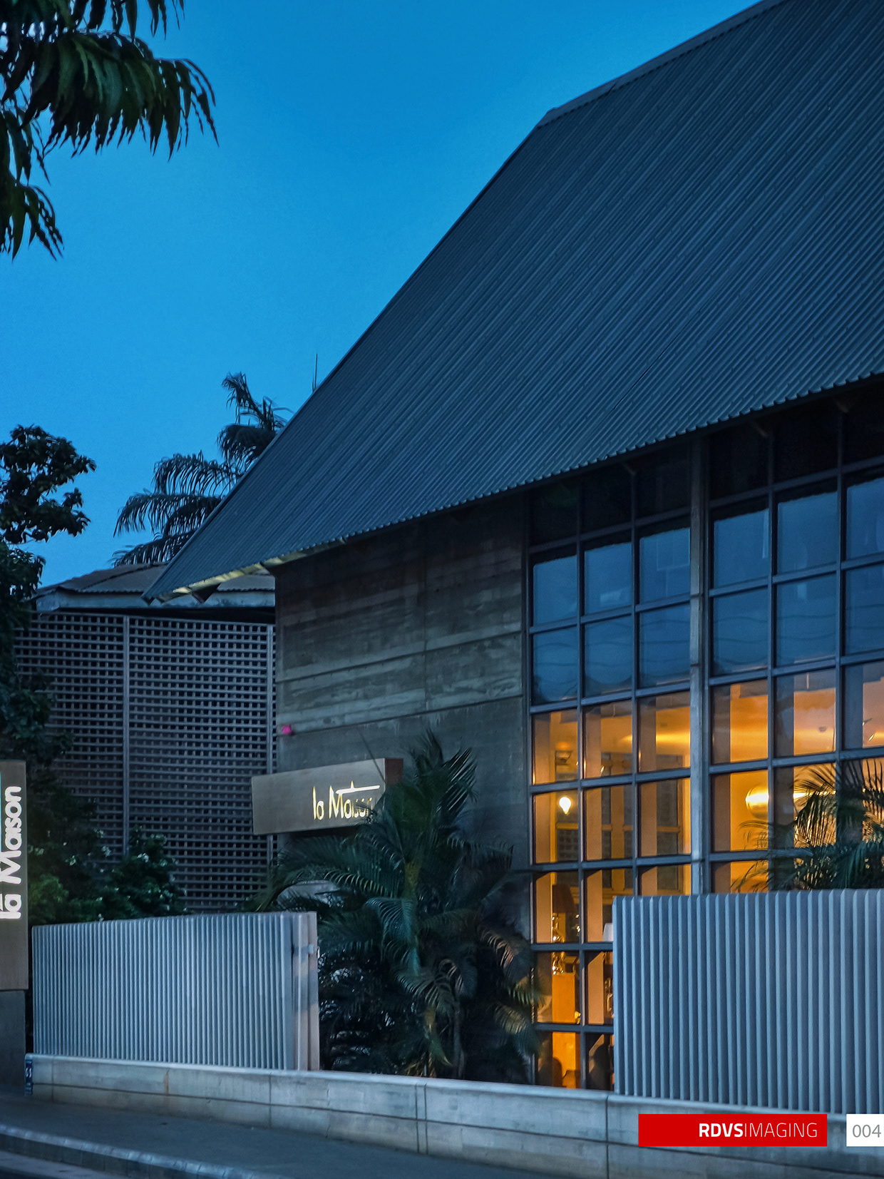 A tall frame looking up at the corner of the building at dusk: the corrugated steel roof plane and its deep open eave cut a hard diagonal from the top left down to the right against saturated blue sky, and palm fronds intrude at the top left and along the base. Below the eave the banded earth-toned wall carries an illuminated blade sign reading la Maison, partly hidden by a small palm, and a vertical sign of the same words is clipped at the far left edge. To the right the full-height steel-framed glazing burns warm gold from the interior lighting, and a pale barred fence on a concrete plinth closes the bottom of the frame, with a perforated lattice screen wall standing behind it at the left. Red RDVSIMAGING badge and a white tag numbered 004 at the bottom right.