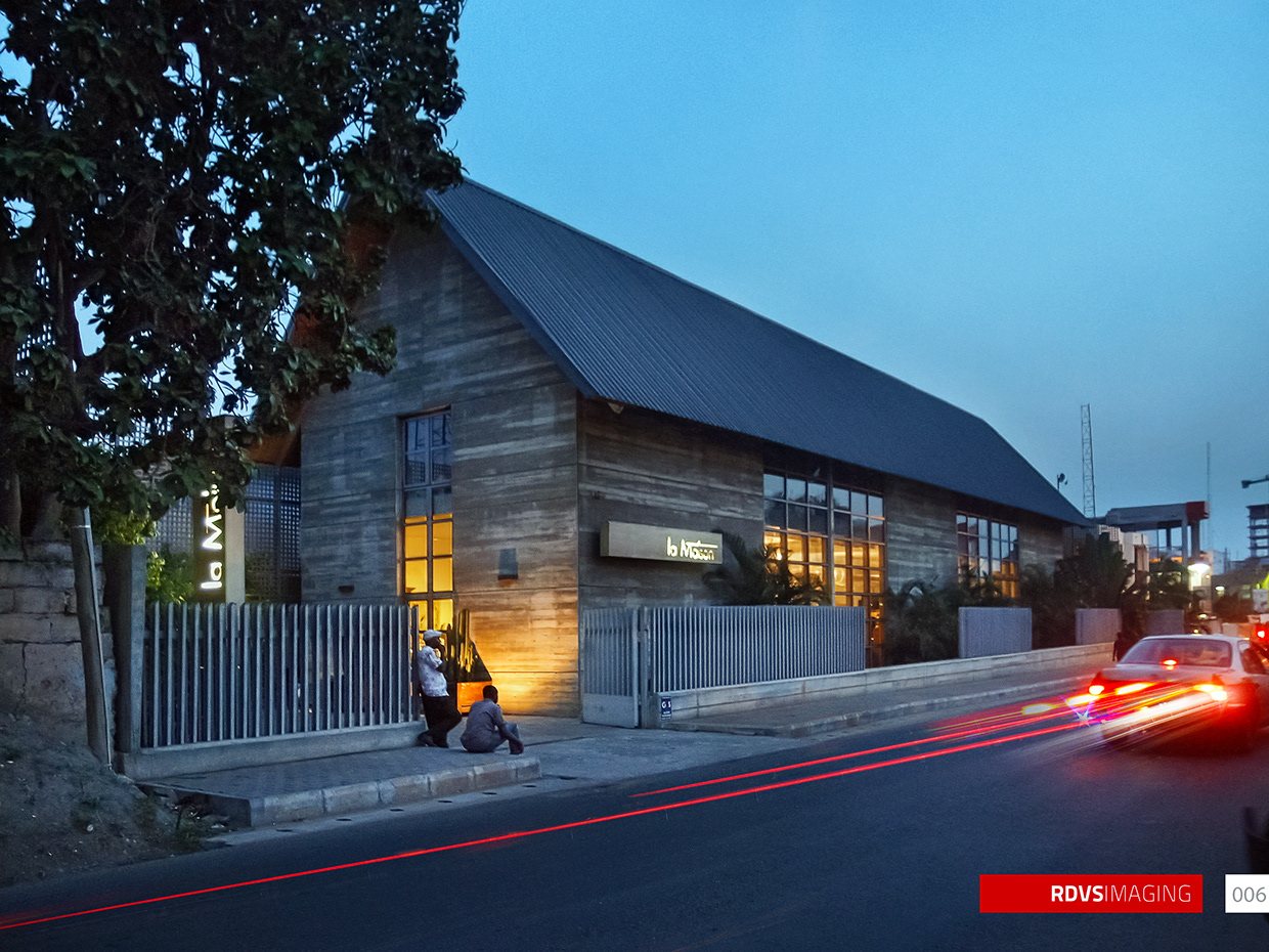 A three-quarter view of the whole building from the road at dusk, made as a long exposure: a car leaving the frame at the right drags three unbroken red tail-light streaks across the asphalt while its headlight flare burns at the kerb. The steep corrugated gable roof runs from the left apex down to the right, the banded earth-toned walls are lit from within by two fields of steel-framed glazing, and the illuminated la Maison blade sign sits at the centre of the frontage with a second vertical sign of the same words at the return. Two men are at the gate on the left, one standing in a pale shirt and one crouched on the kerb, behind a pale barred fence; a leafy tree fills the top left corner and a distant crane and unfinished building stand at the right. Red RDVSIMAGING badge and a white tag numbered 006 at the bottom right.
