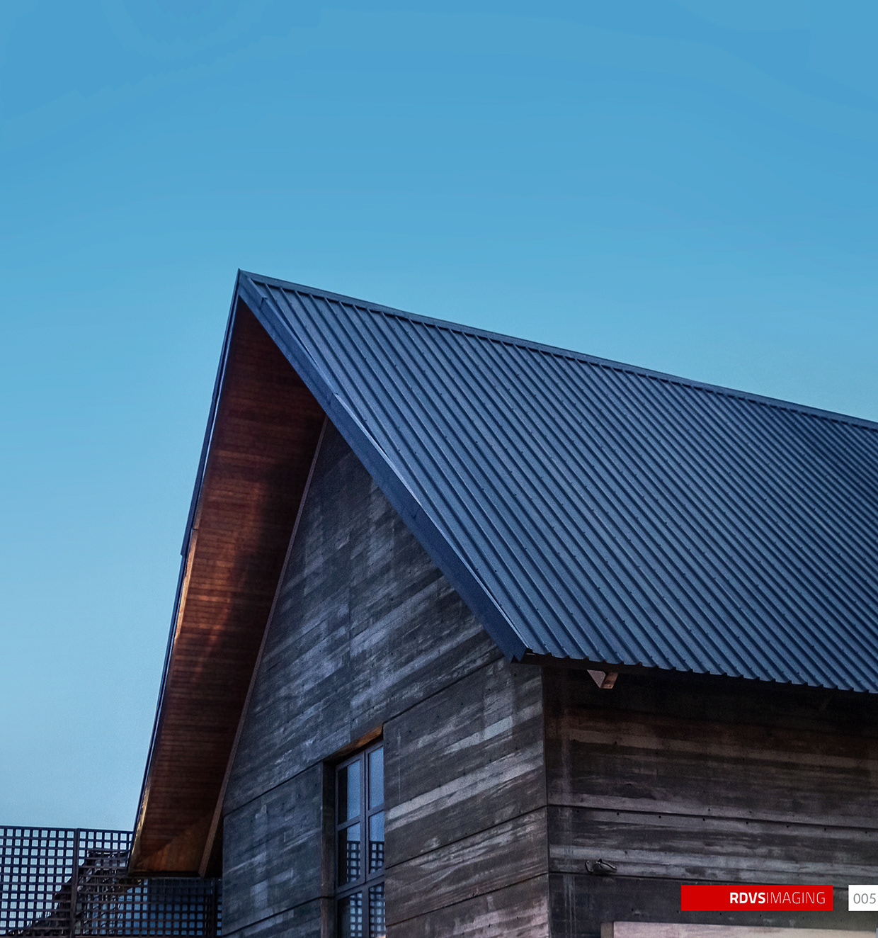 A near-square detail of the gable apex against clear blue dusk sky: the corrugated steel roof planes meet at a sharp apex in the upper left, and the underside of the deep overhang is lined in warm-toned boards that catch the last of the light. Below the eave the banded earth-toned wall shows its horizontal shuttering marks and a scatter of tie holes, with a small steel-framed window set high in it and a bracket light fixed lower down. A strip of perforated lattice screen is visible at the bottom left and the edge of the blade sign along the bottom right. Red RDVSIMAGING badge and a white tag numbered 005 at the bottom right.