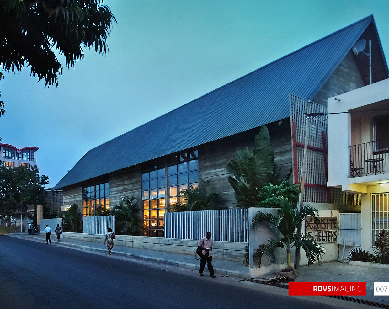 The long street elevation seen from the far end at dusk: the corrugated gable roof runs away to the right, where a satellite dish is fixed to the gable end, and a white two-storey neighbouring house with a balcony and a hand-painted board reading XQUISITE SHELTERS, its leading letter gone at the edge of the board, stands at the right edge. The banded earth-toned wall is fronted by a pale barred fence on a low concrete plinth with two gated vehicle openings, and two fields of full-height steel-framed glazing glow warm at the near end. Banana plants and palms fill the compound behind the fence, a tall narrow sign panel stands unreadable at the far left, and four pedestrians are on the pavement in front, one in a pink shirt crossing the centre carrying a bag. A red-roofed tower shows at the far left. Red RDVSIMAGING badge and a white tag numbered 007 at the bottom right.