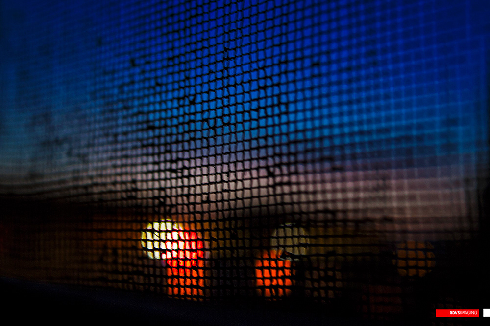 A window screen held close and tilted, so the woven fibreglass mesh fills the whole frame. The grid is tack-sharp only in a band across the middle and upper right, where the squares read as a clean dark lattice against blue, and it dissolves into a soft moire at the left edge and across the top. Behind the mesh the upper half is a deep blue dusk sky grading to a dull band of cloud, while the lower half is near black with out-of-focus lights burning through: a bright white-yellow orb left of centre with red-orange smears hanging below it and a second, dimmer disc to its right. The only close-up and the only frame whose subject is a texture rather than a view. Red RDVSIMAGING badge, bottom right.