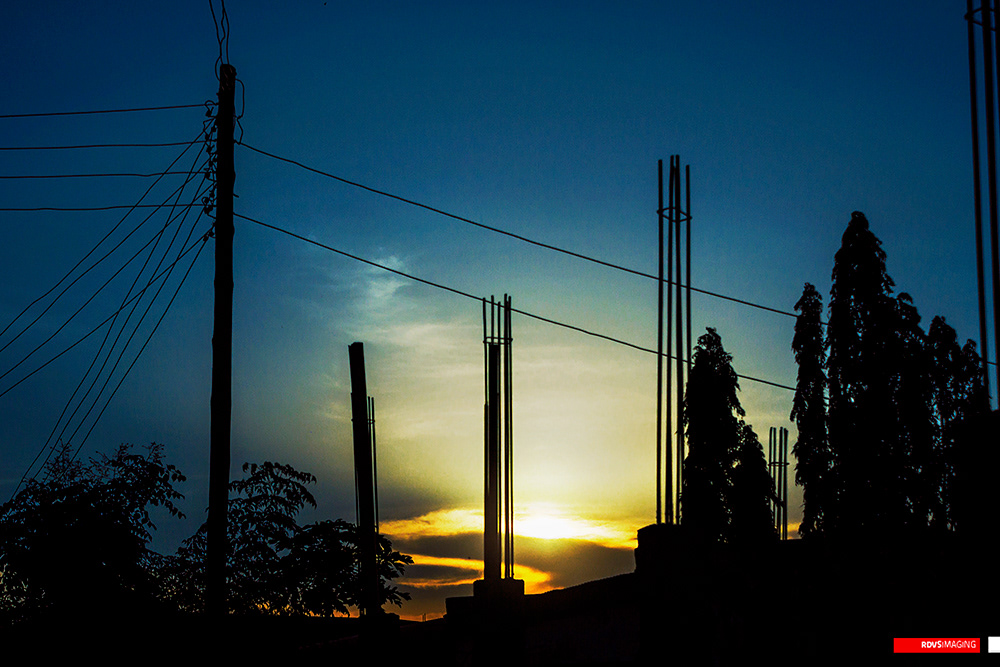 Immediately after sunset: a tall blue sky with a thin lens of cloud low down, lit from underneath, and the sun reduced to a bright slit behind a black roofline. A tall wooden utility pole stands at the left with a fan of service drops running out of its crossarm and off the top-left corner of the frame, and a single cable spans the whole width from it to the right. Three concrete column starters with their crowns of exposed rebar rise at different heights across the middle, a leafy shrub sits at the lower left, and a stand of narrow trees fills the right. Red RDVSIMAGING badge, bottom right.