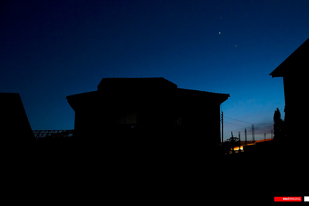 Almost entirely silhouette: a deep blue post-sunset sky, darkest at the top, with three small points of light in the upper right. A two-storey house with a hipped roof and a wide eave stands black and unreadable in the centre, its only detail a faint rectangle of louvered shutter. The corner of a roof cuts in at the left with a lattice of roof trusses or a solar frame below it, and the gable end of a third building closes the right edge. Between the house and that gable the horizon survives as a narrow slot of orange afterglow, in front of which stand a line of rebar-crowned prods, a utility pole and a single tree. Red RDVSIMAGING badge, bottom right.