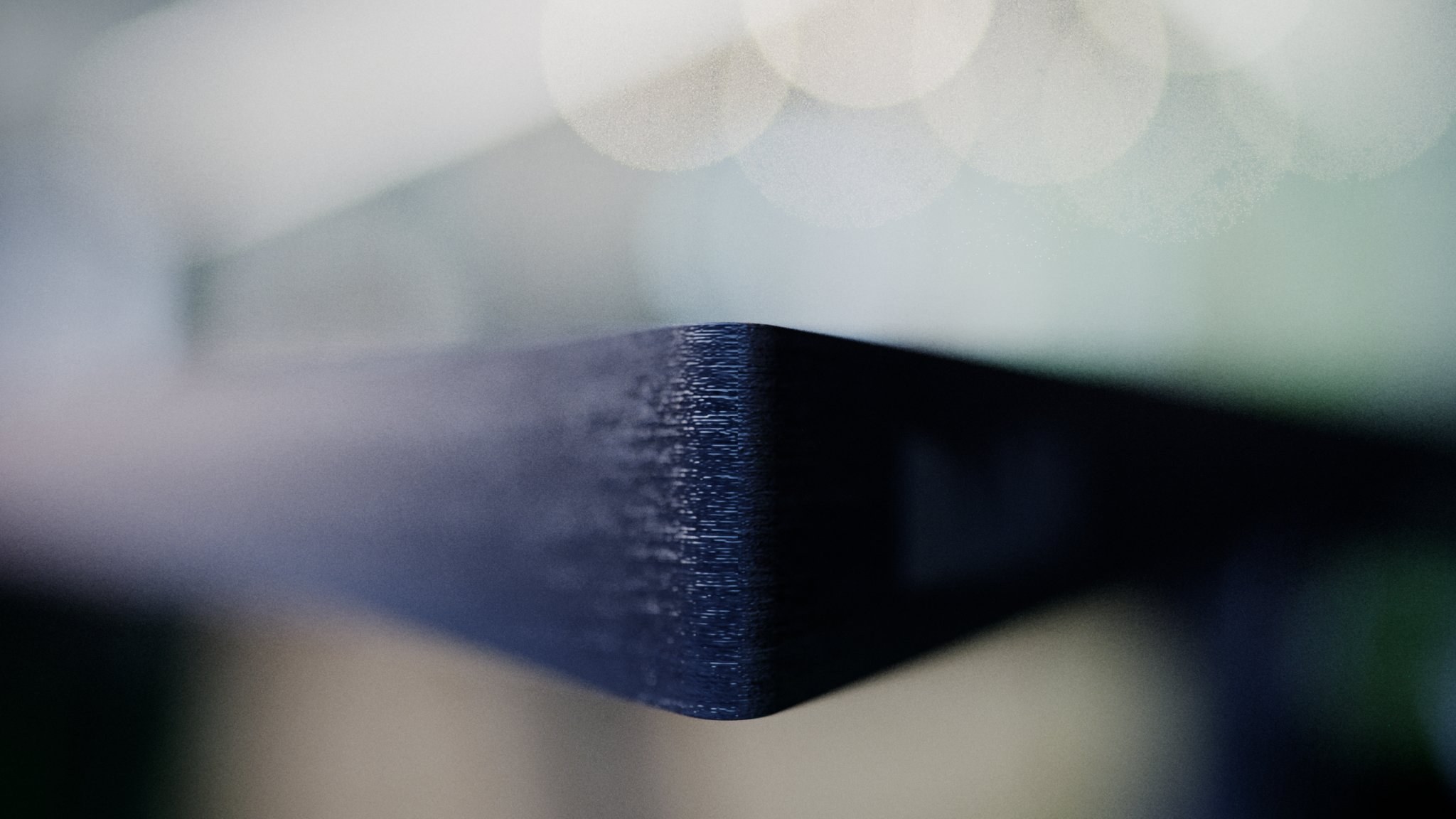 MIG HG-DESK macro at the tip of a rail: a navy-black stained wood edge with fine open grain and a rounded vertical corner held in sharp focus against pale bokeh discs.