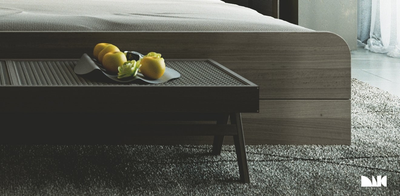 MIG MOUND in a bedroom at floor level: the same grey tray of yellow fruit and green flowers set on a dark wood bench with a perforated top at the foot of a made bed, grey loop-pile rug and sheer curtains at right.