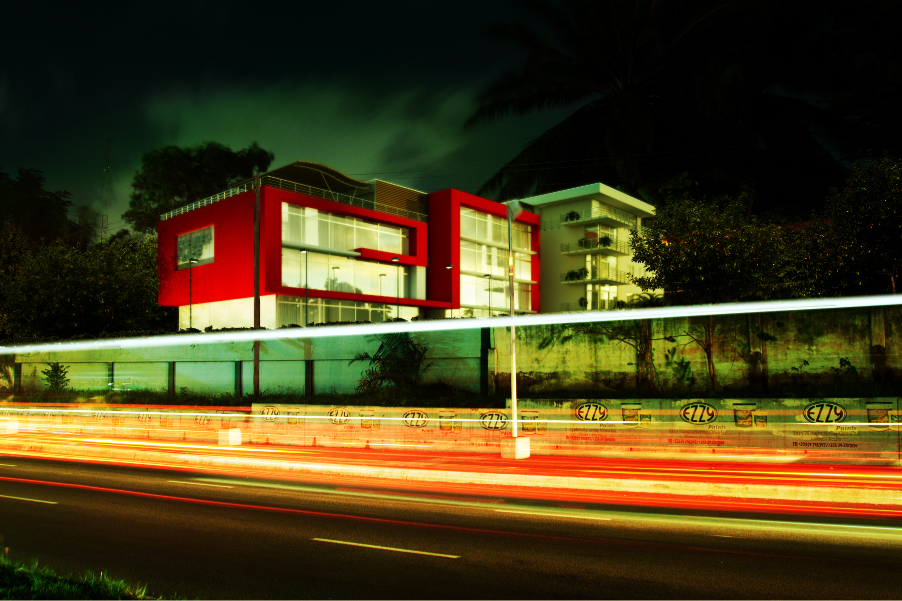 Premier Lodge 2 at night, long-exposed from across the highway: a red-framed glass volume lit from within beside a pale balconied apartment block, a painted perimeter wall behind the traffic and white and red light trails streaking the foreground