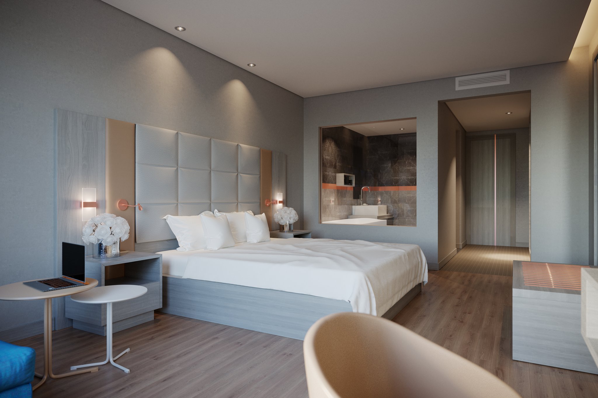 Protea Hotel Airport king room at dusk: a made bed against a padded square headboard with copper reading arms and sconces, vases of white flowers on the nightstands, a laptop on a small round table, and a glazed opening onto the stone bathroom.