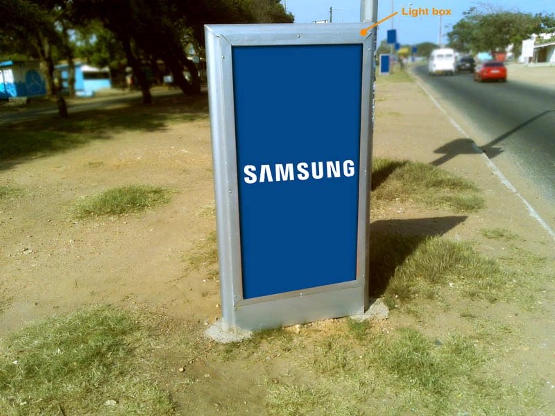 Photograph of a totem light box installed on a grassy roadside verge, blue face with white SAMSUNG lettering, labeled Light box in orange, traffic and trees receding along the highway.